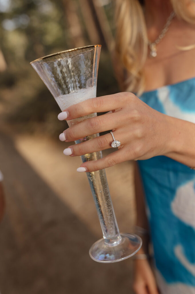 Close-up of an oval engagement ring on a woman’s hand as she holds a tall champagne flute against her blue patterned dress