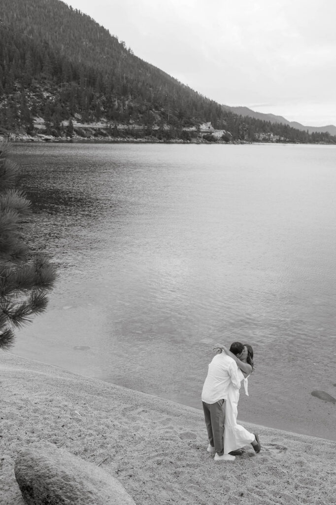 Black-and-white landscape of an engaged couple kissing beside the calm water and mountains of Lake Tahoe