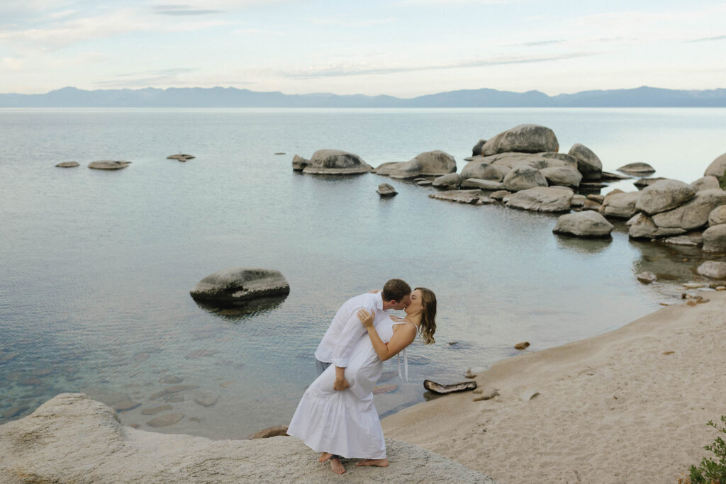 Engaged couple sharing a dip kiss on the sandy shoreline during sunrise at Lake Tahoe