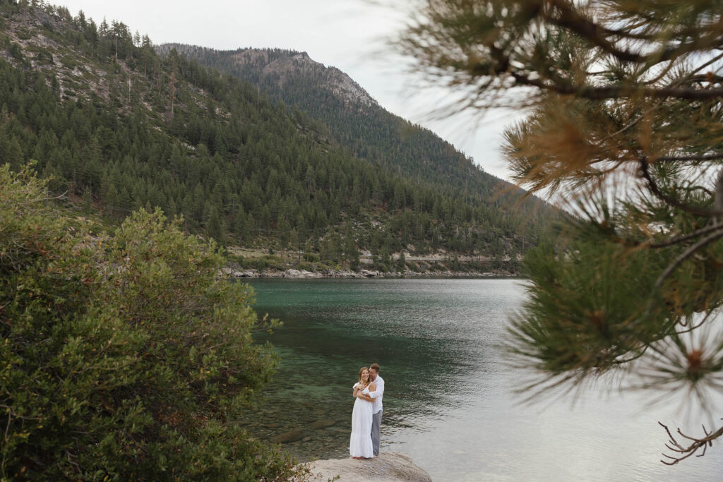 Sunrise Lake Tahoe engagement photo of a couple framed by pine trees, mountains, and emerald water