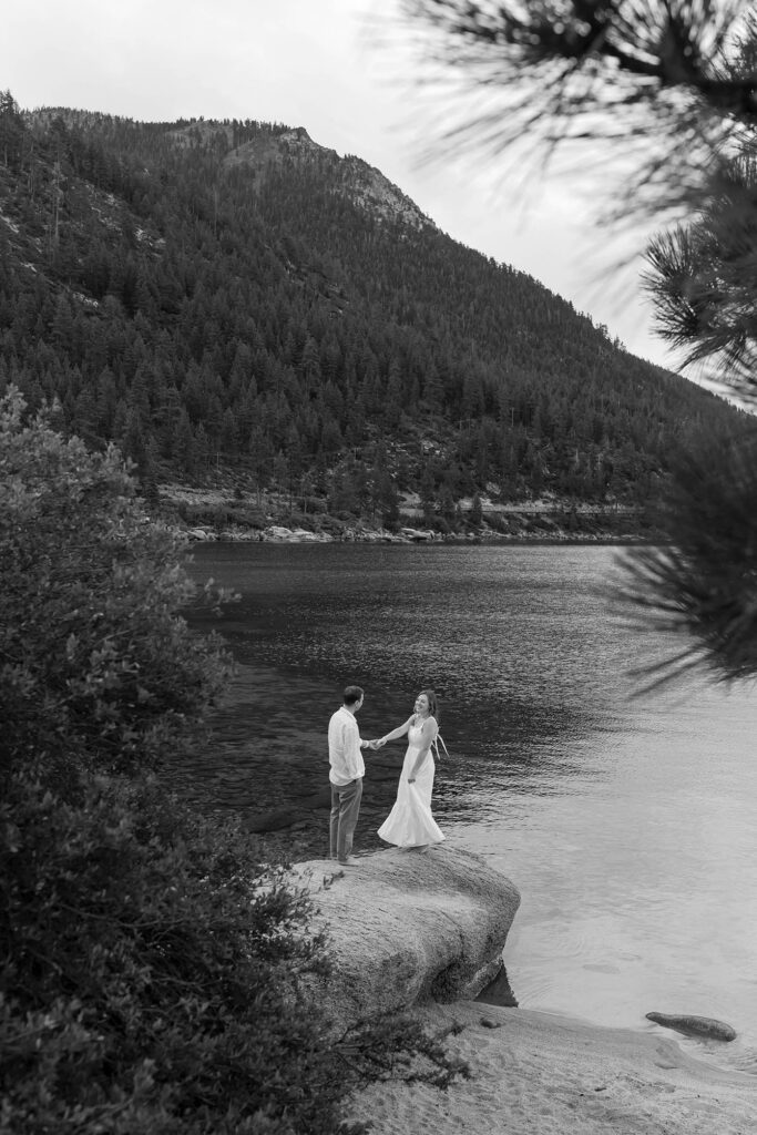 Black-and-white landscape of a couple holding hands on a rocky ledge above Lake Tahoe