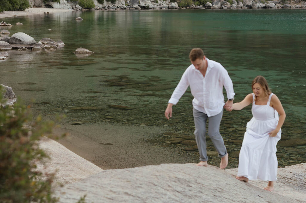 Engaged couple walking barefoot hand in hand across a granite rock beside the lake