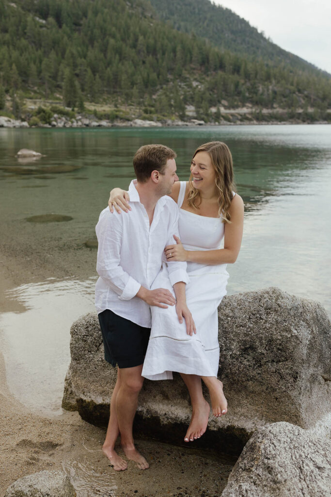 Engaged couple sitting barefoot together on a granite boulder beside the clear water of Lake Tahoe