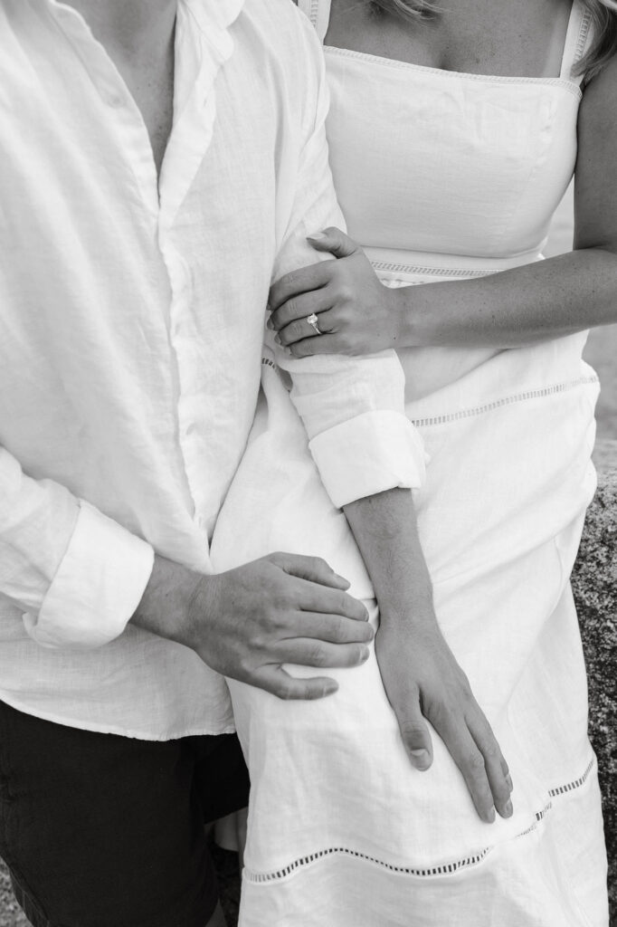 Close up black-and-white photo of a woman's engagement ring as she holds her fiancé’s arm
