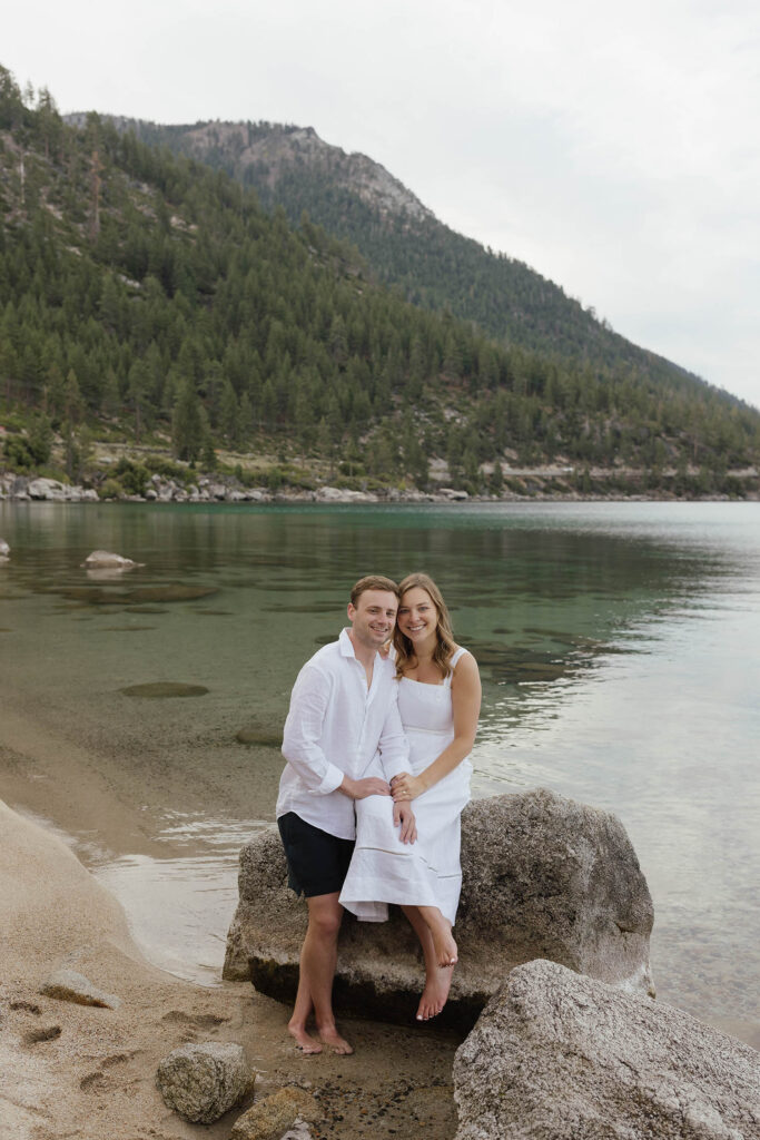Barefoot, smiling couple sitting on a rock together with Lake Tahoe's aqua water and mountains behind them