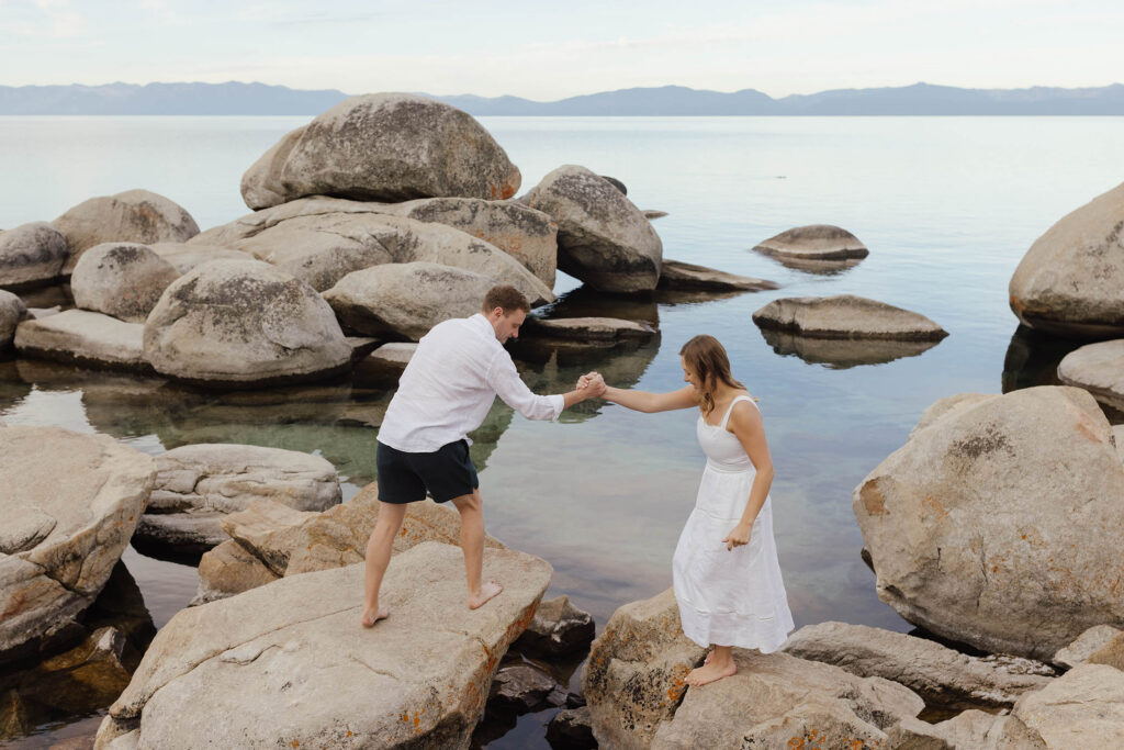 Man helping woman in white dress across granite boulders along the Lake Tahoe shoreline for their engagement photos