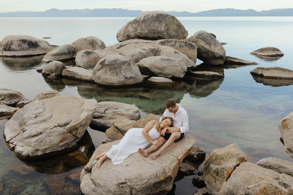 woman lays in mans lap on large granite boulders in Lake Tahoe while he runs his fingers through her hair for their engagement photos