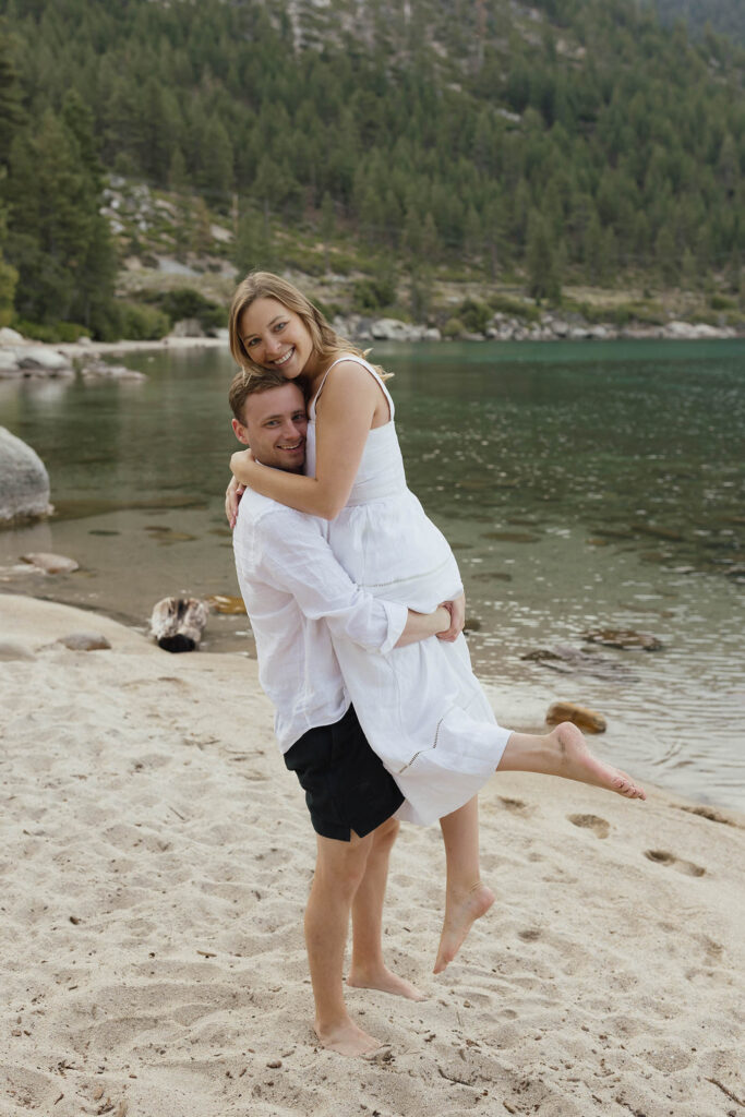 Man lifts up woman while they smile at the camera and she pops one foot on the beach in Lake Tahoe