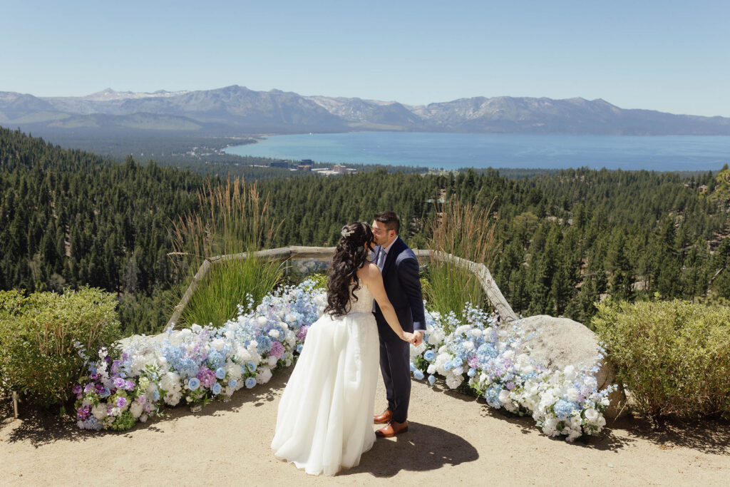 Bride and groom kissing beside blue and lavender flowers with a panoramic Lake Tahoe view