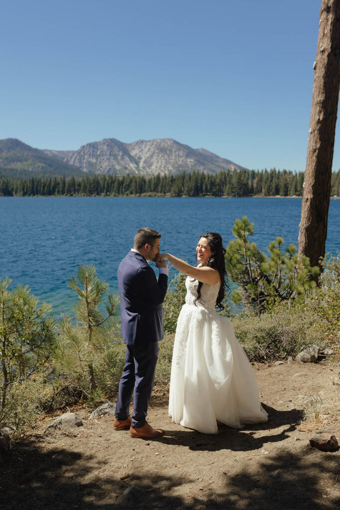 Groom kissing the bride’s hand beside a blue alpine lake surrounded by pine trees and mountains
