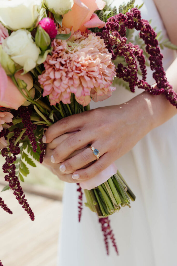 Close-up of the bride’s blue gemstone engagement ring as she holds her colorful wedding bouquet