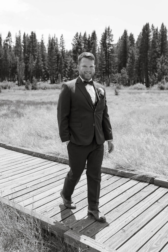 Black-and-white portrait of the smiling groom standing on a wooden boardwalk with pine trees behind him