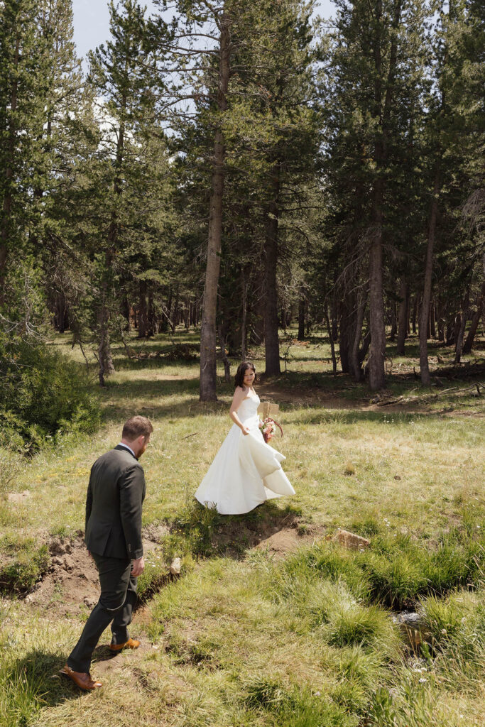Bride lifting her dress and looking back as the groom follows her through a grassy clearing among Lake Tahoe pine trees