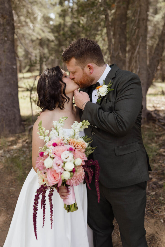Bride and groom kissing in a pine forest, with the bride holding a cascading pink, white, and burgundy bouquet