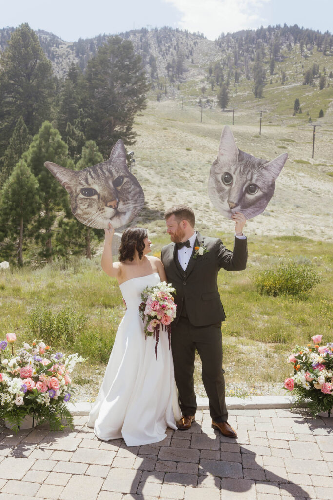 Bride and groom holding oversized cat face cutouts at an outdoor wedding ceremony site overlooking grassy ski slopes