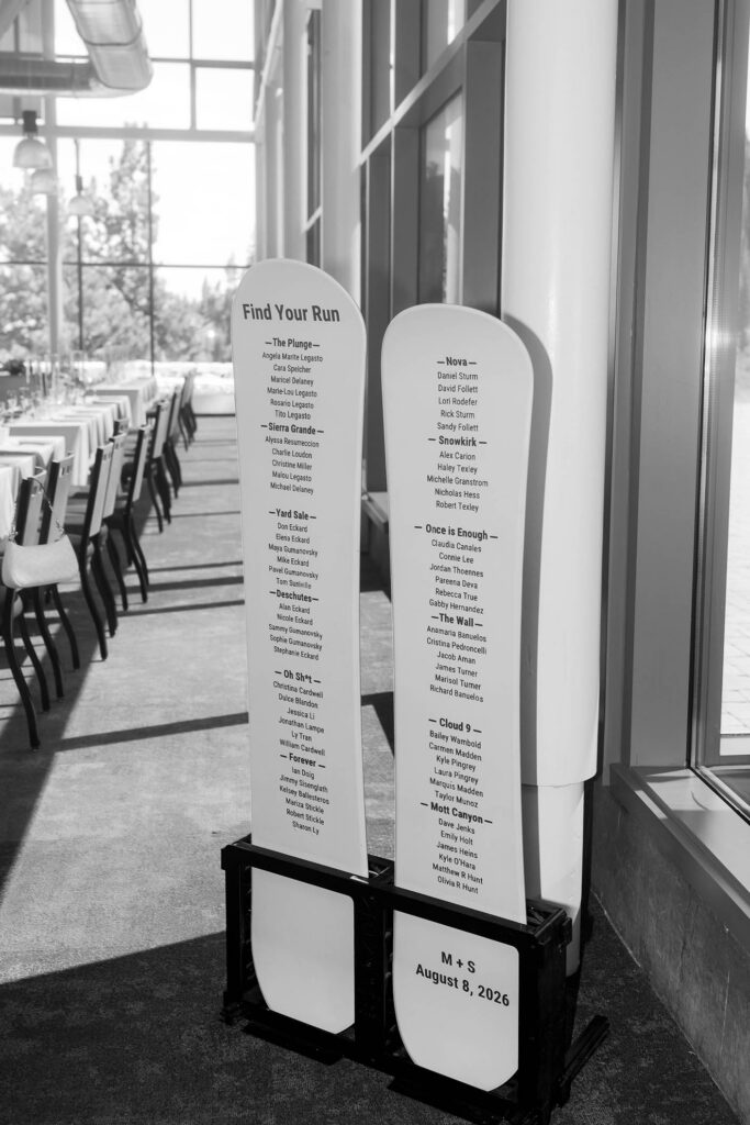 Black and white photo of a ski-shaped wedding seating chart labeled Find Your Run beside tall reception windows