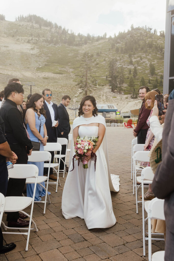 Bride wearing a flowing white cape and carrying a colorful bouquet as she walks down the aisle at Winters Creek Lodge