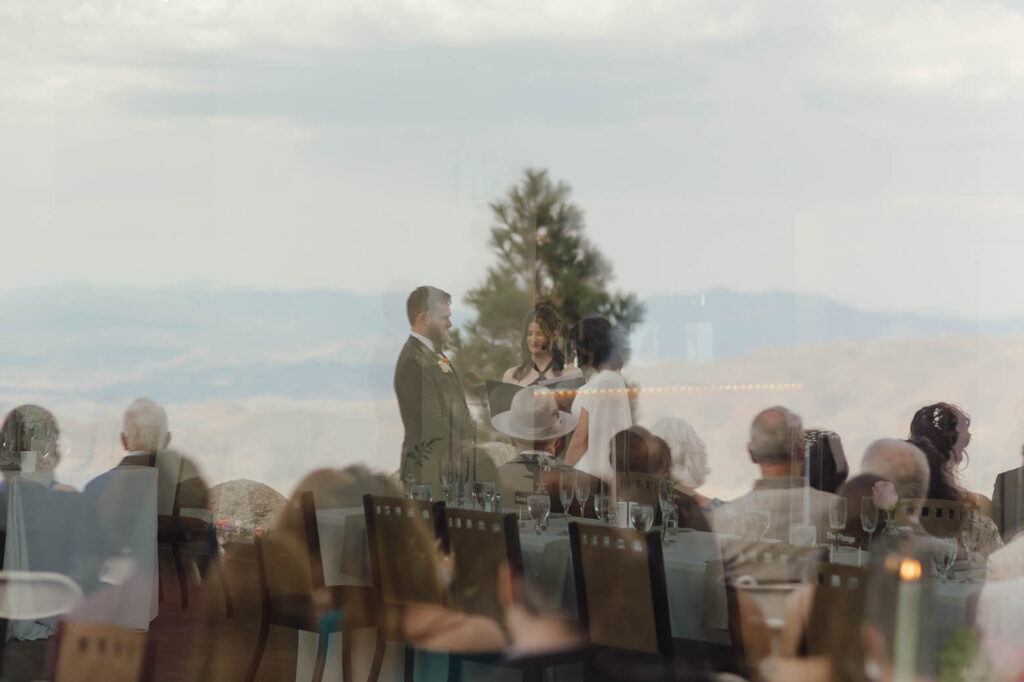 Bride and groom during their Winters Creek Lodge ceremony, photographed through glass with reflections of reception tables