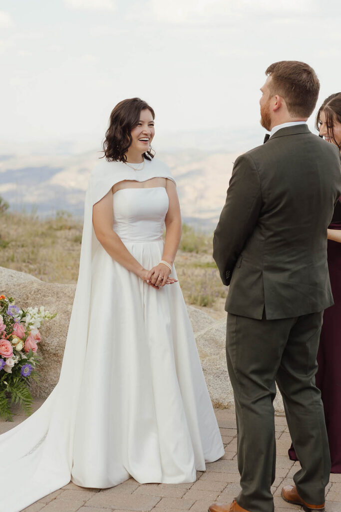 Bride laughing with the groom during their outdoor ceremony, wearing a white gown and cape with mountains behind her