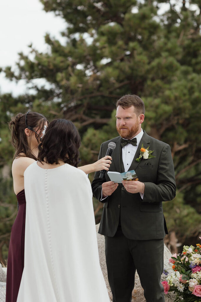 Groom reading his vows from a booklet while the bride listens and the officiant holds a microphone at Winters Creek Lodge