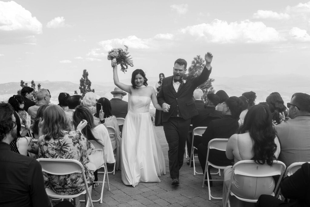 Black and white photo of the bride and groom raising their arms while walking down the aisle as guests applaud
