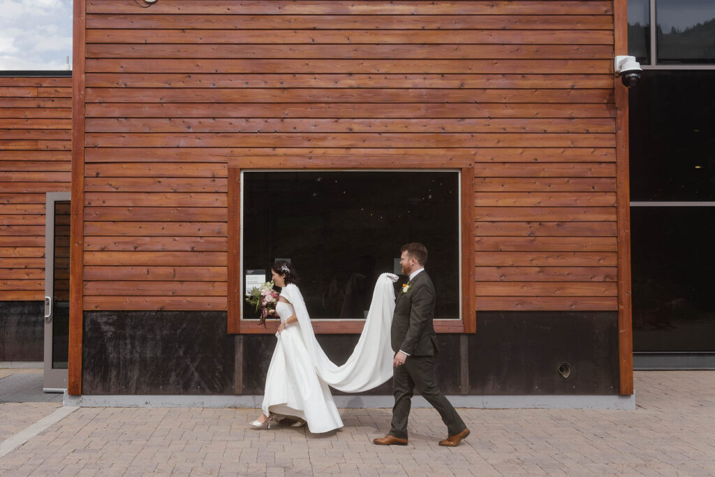 Groom carrying the bride’s flowing white cape as they walk beside the wood exterior of Winters Creek Lodge