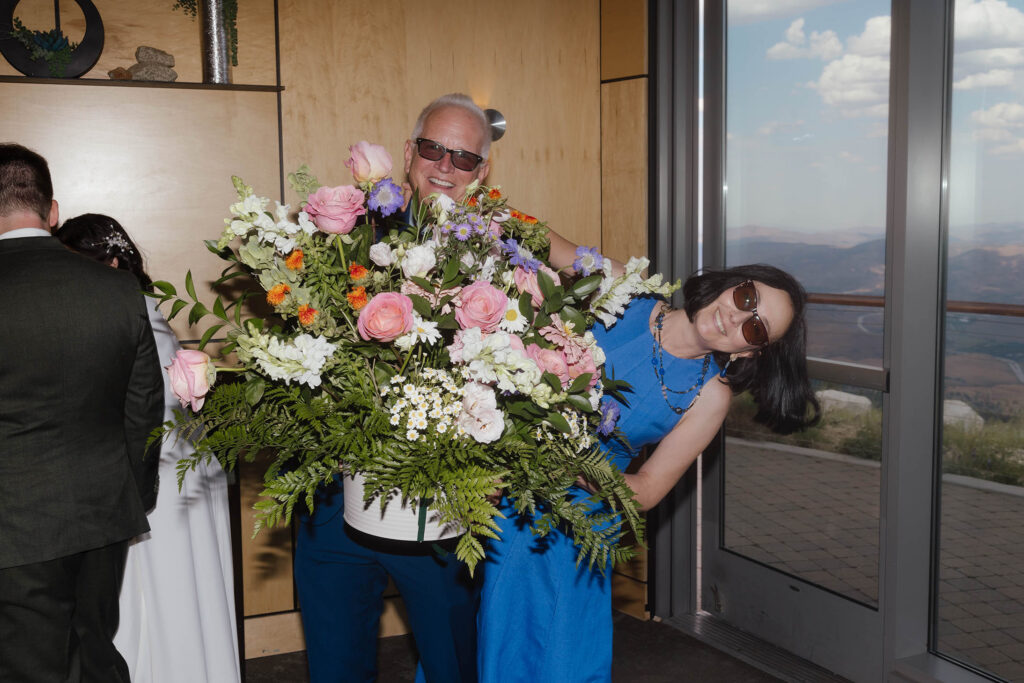 Two wedding guests in blue smiling behind a large arrangement of pink, white, purple, and orange flowers inside Winters Creek Lodge