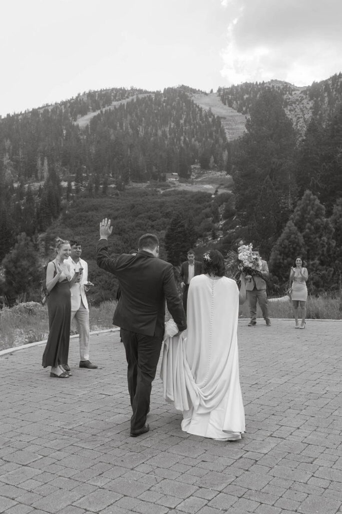 Black and white photo of the bride and groom greeting guests outside Winters Creek Lodge with forested ski slopes behind them