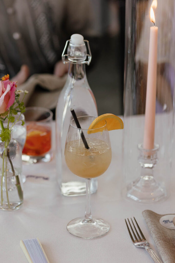 Cocktail garnished with an orange slice beside a pink taper candle and flowers on a wedding reception table