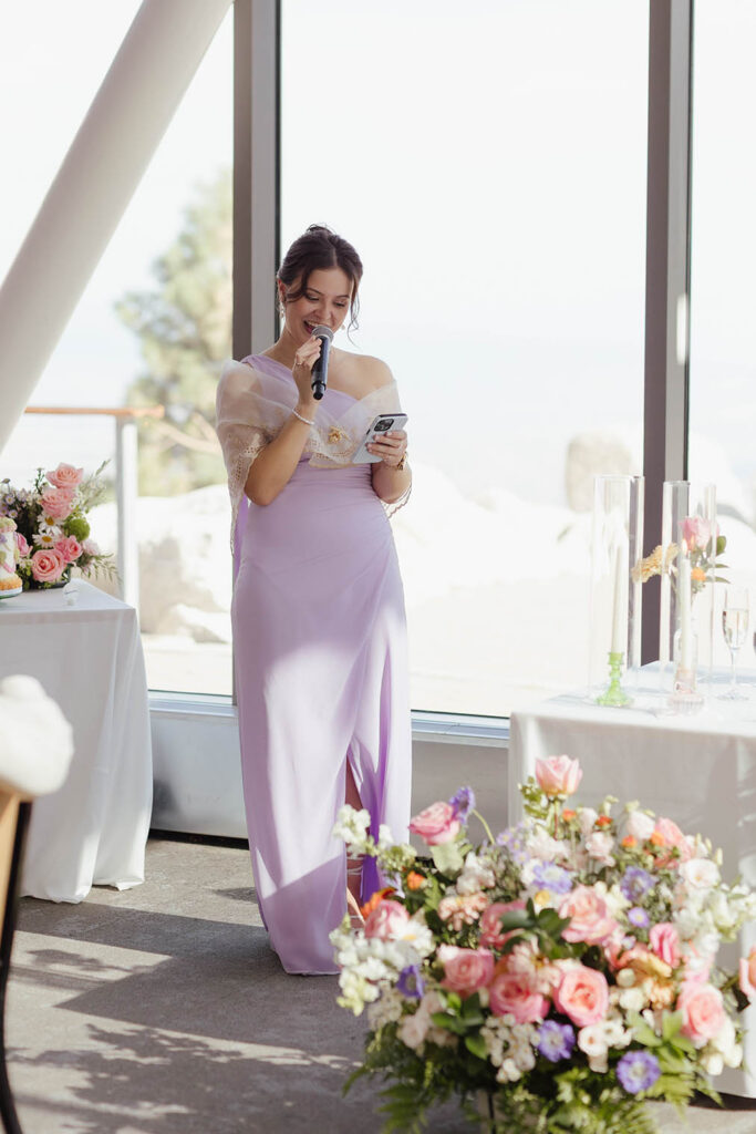 Wedding guest in a lavender dress reading a speech from her phone beside colorful flowers at Winters Creek Lodge