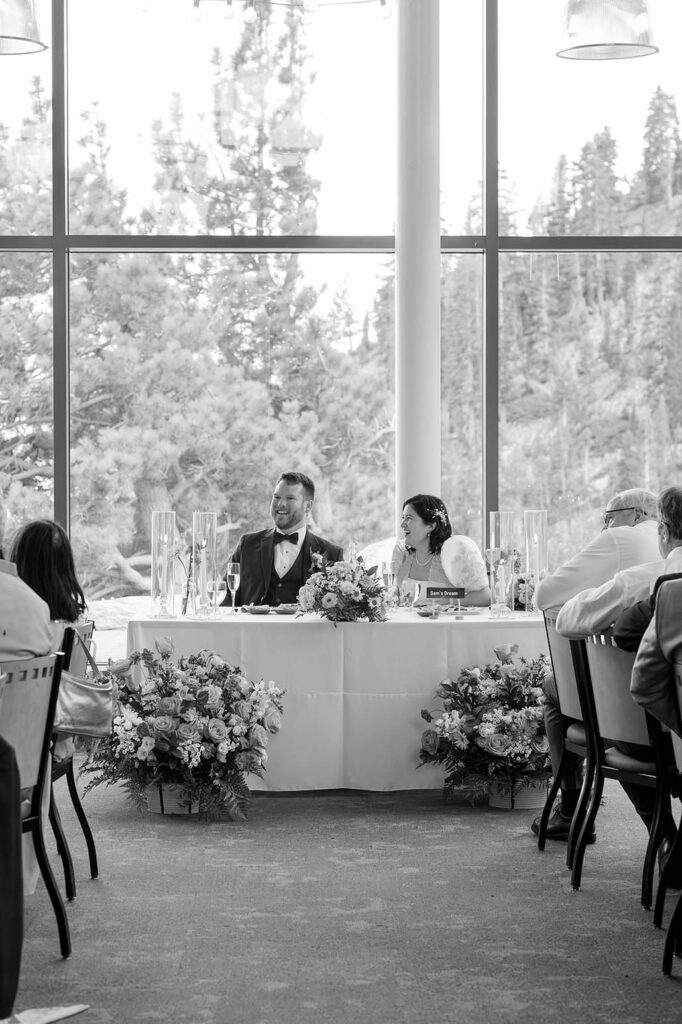 Black and white photo of the bride and groom at their sweetheart table beneath tall windows overlooking pine trees