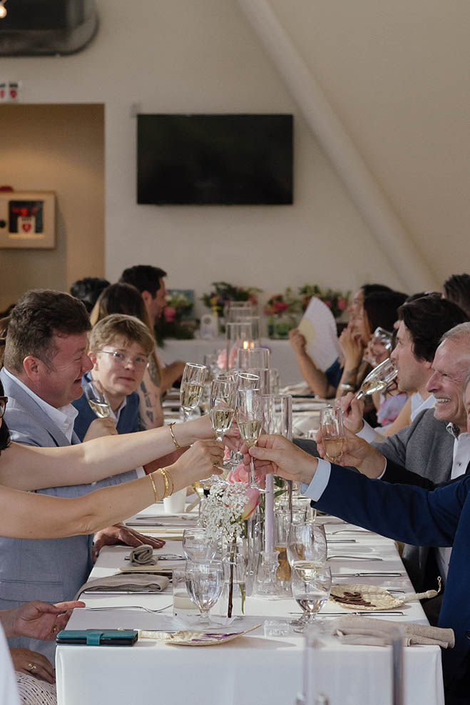 Wedding guests clinking champagne glasses across a reception table decorated with flowers and pink taper candles