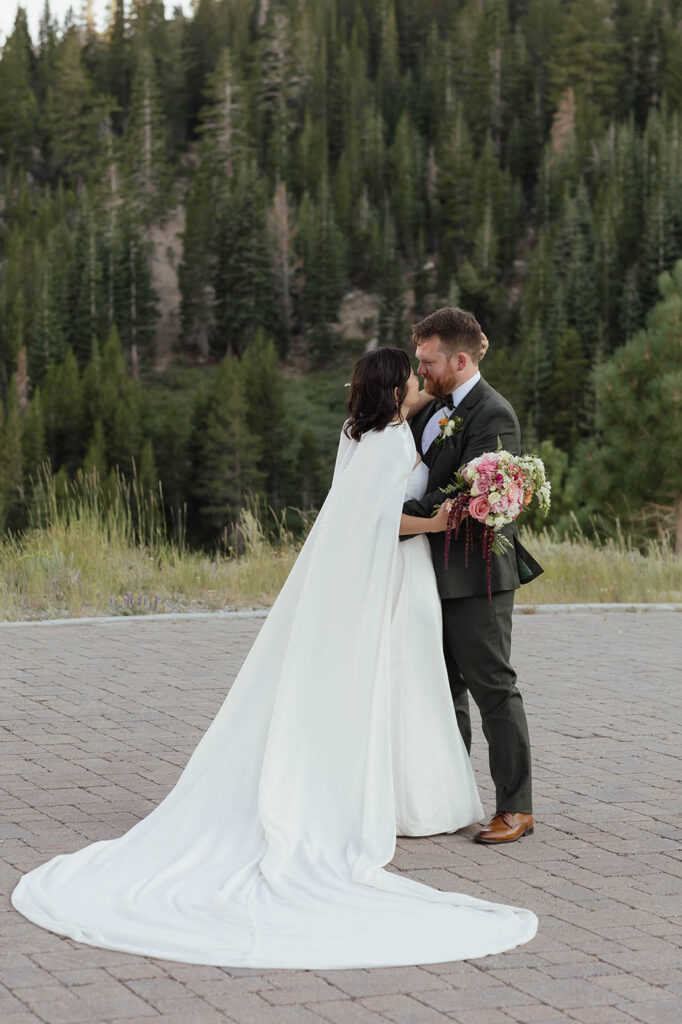Bride and groom embracing on the Winters Creek Lodge terrace with the bride’s long white cape spread across the pavers