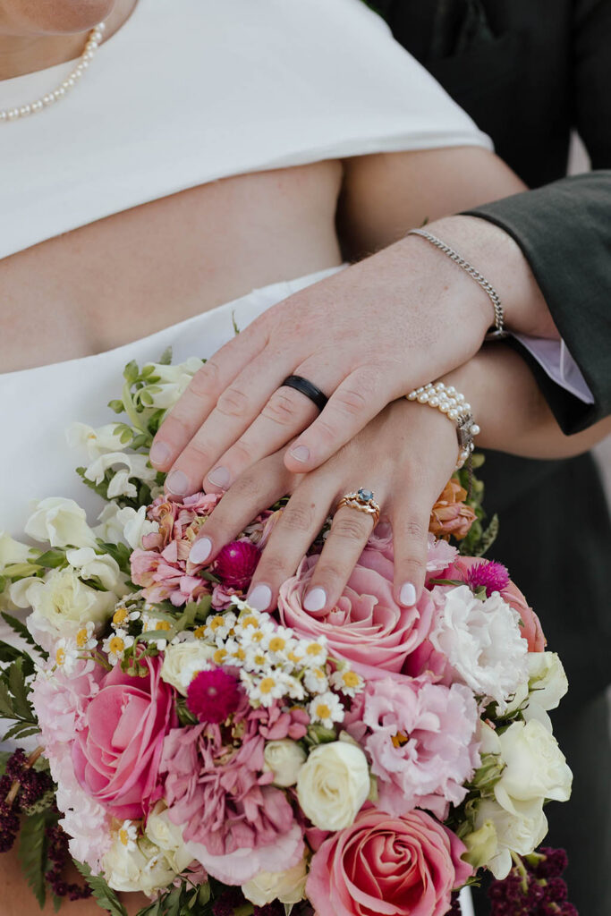 Close-up of the bride and groom’s wedding rings as they rest their hands over a pink and white bridal bouquet