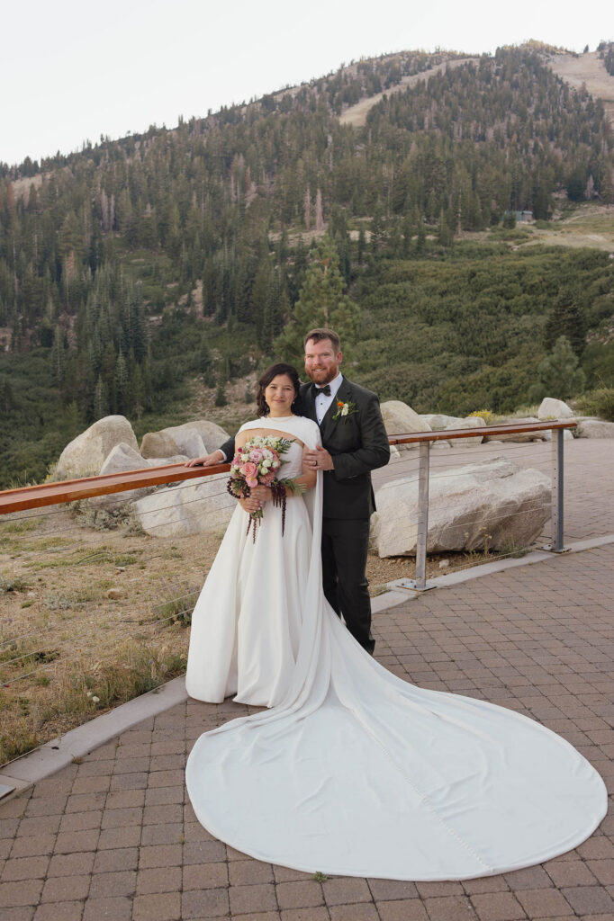 Bride and groom posing beside the terrace railing at Winters Creek Lodge with a sweeping white cape and forested mountain backdrop