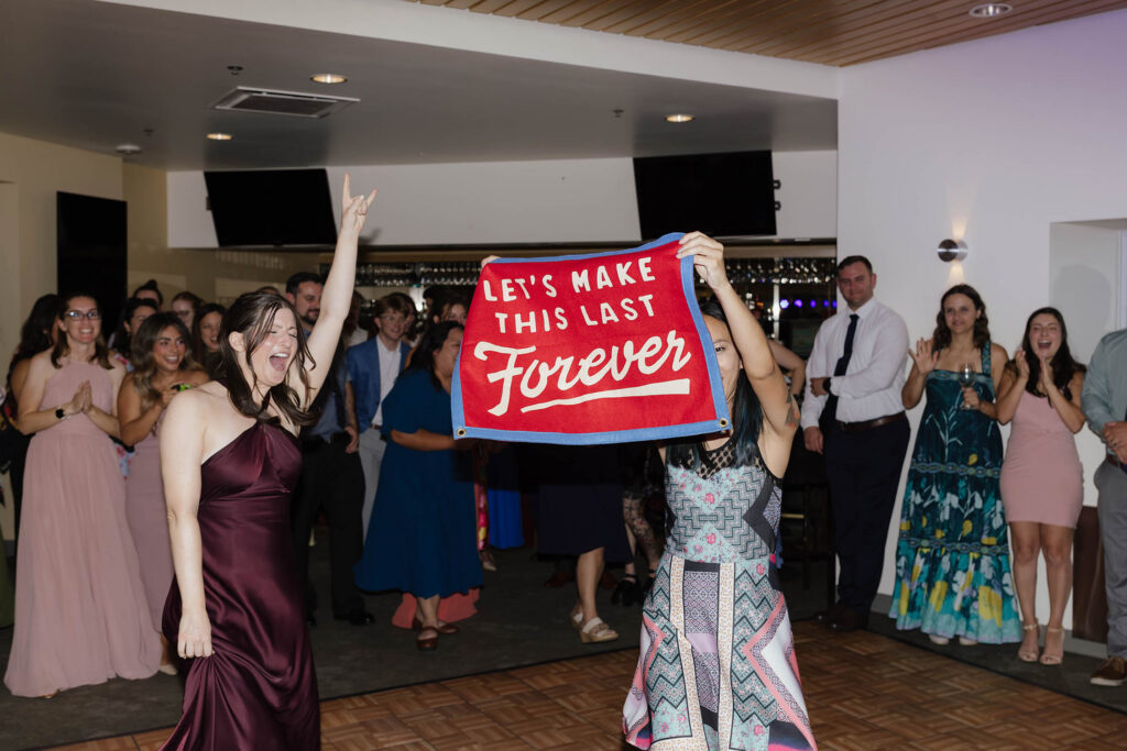 Wedding guests cheering on the dance floor while holding a red banner reading Let’s Make This Last Forever