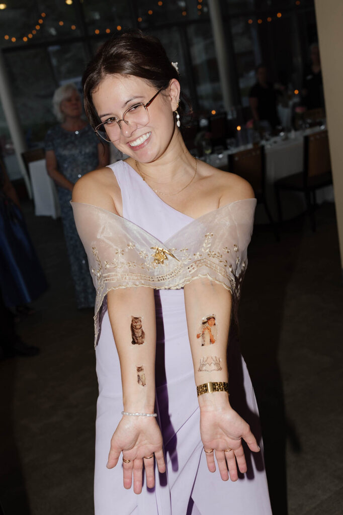 Smiling wedding guest in a lavender dress showing playful temporary tattoos on both forearms