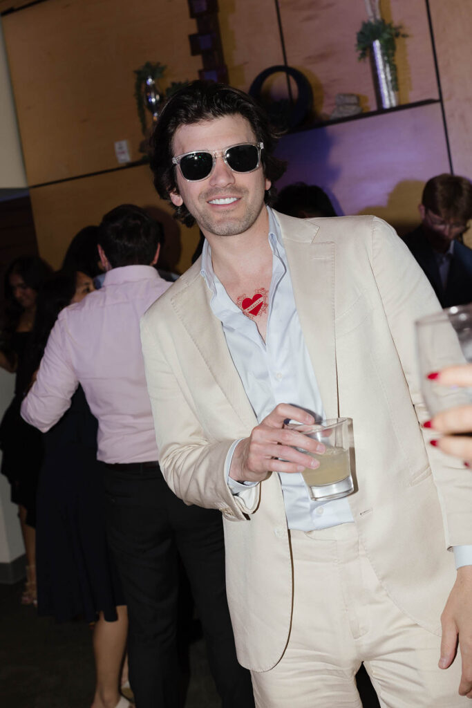 Wedding guest in a cream suit and sunglasses holding a cocktail and showing a red heart temporary tattoo